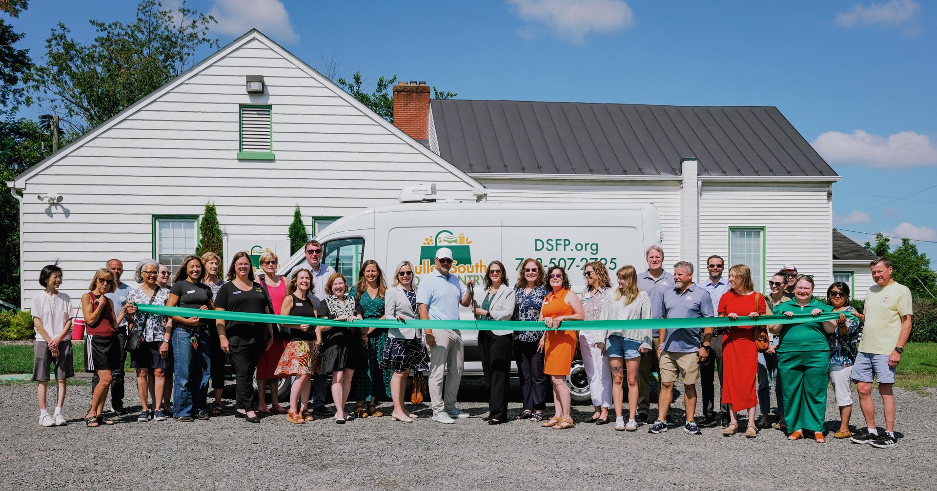Large group of people standing outside in front of the Pantry and new cargo van holding a green ribbon
