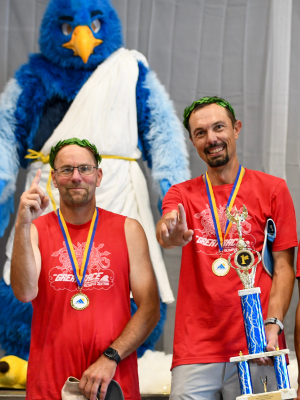 Two individuals wearing matching red athletic shirts and black shorts stand on a stage holding medals and a large trophy. They each have green laurel wreaths on their heads and are displaying their awards.