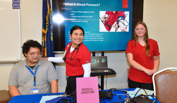 Three CEI students stand at a table labeled “Medical Assisting” with blood pressure equipment, in front of a screen explaining blood pressure.
