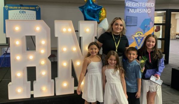 Caitlyn Keelin stands with her four children in front of large, illuminated “RN” letters. Behind them are blue and gold balloons and a banner displaying the words “Registered Nursing.”