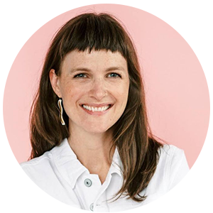 Smiling headshot of person with long, dark brown hair wearing white and gold dangle earrings and a white, collared top. The background of the photo is pink.