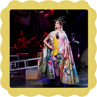 Person in colorful, graffiti-style cape poses on stage with musicians and dramatic lighting.