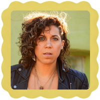 A photo of musician Kristen Ford. Kristen has curly brown, shoulder length hair, is wearing a black leather jacket and layered necklaces. 