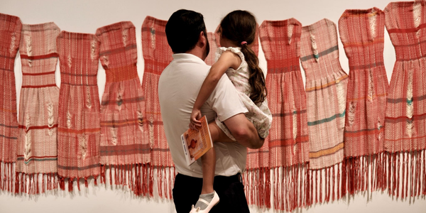 A caretaker holding a young child stands in front of a display of traditional woven dresses with red patterns and fringes hanging on a wall in an exhibition or museum.