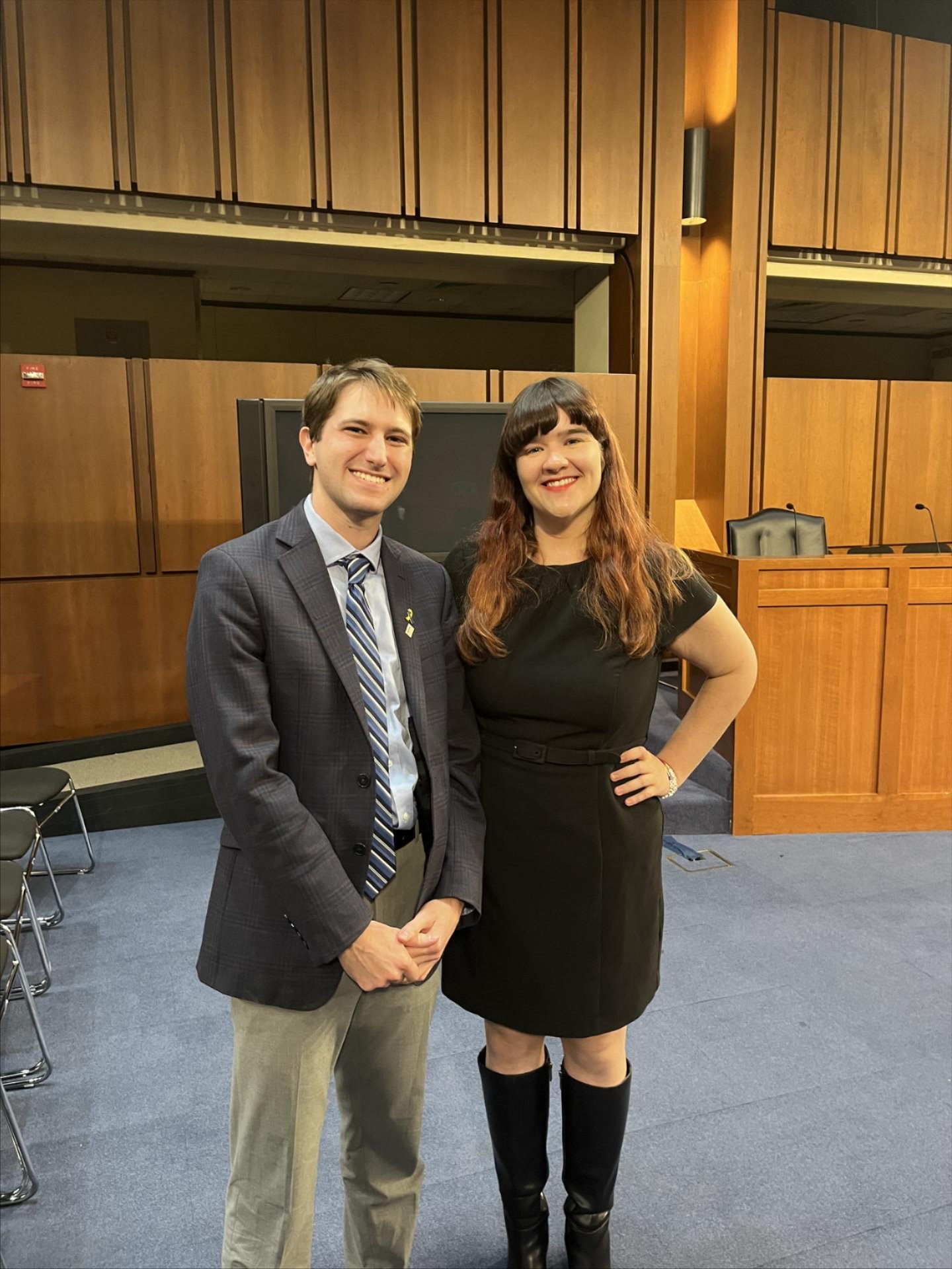 Micah Rothkopf, a white man with short brown hair wearing a suit, smiles next to Grace Hart, a woman with long dark hair with a red gradient, wearing a black dress and black boots, and are standing together in a Senate hearing room.