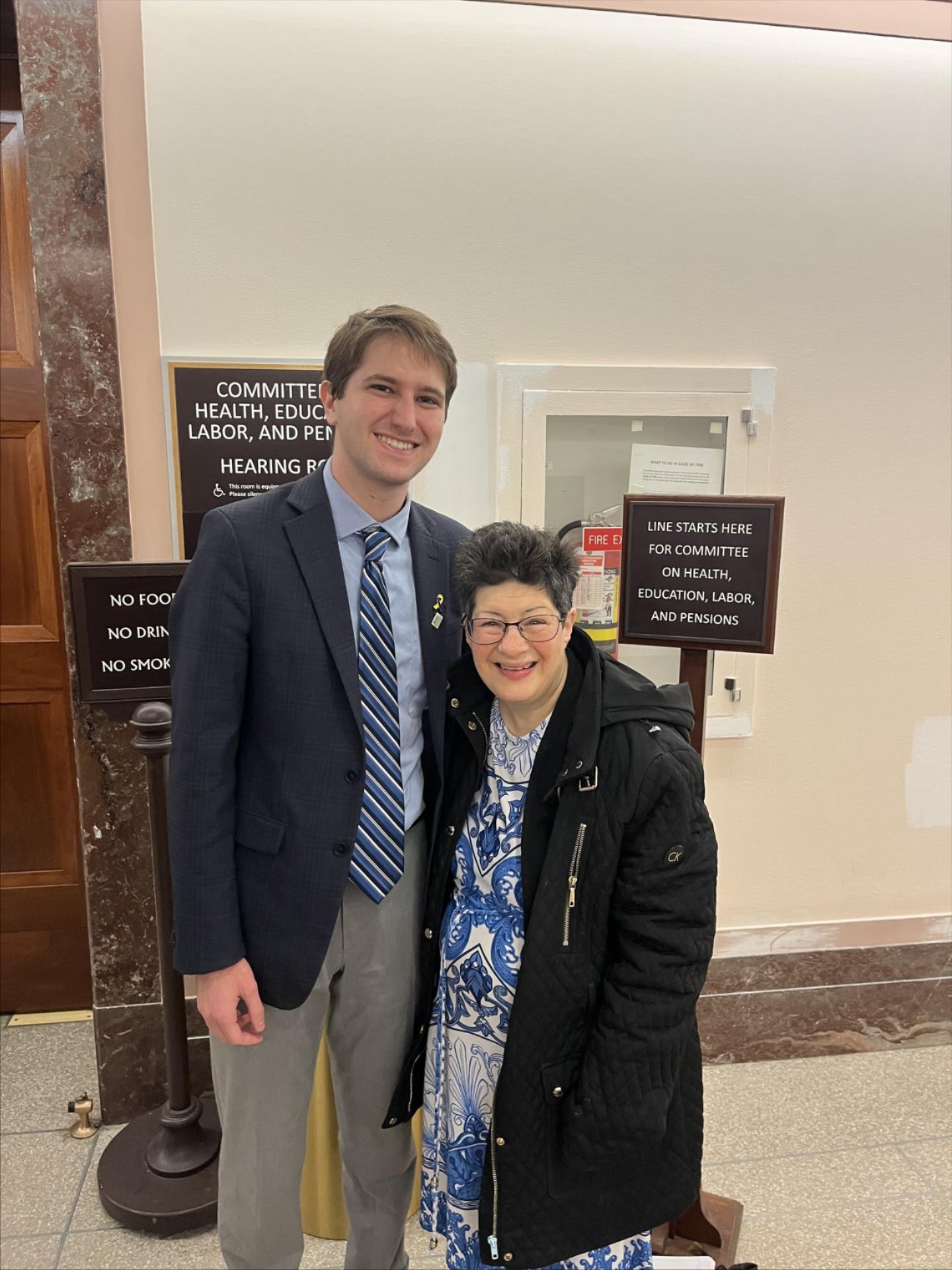 Micah Rothkopf, a white man with short brown hair wearing a suit, smiles next to Liz Weintraub, a woman wearing glasses, a jacket, and a blue and white patterned dress, and are standing together in front of a sign for the Senate HELP Committee hearing room.