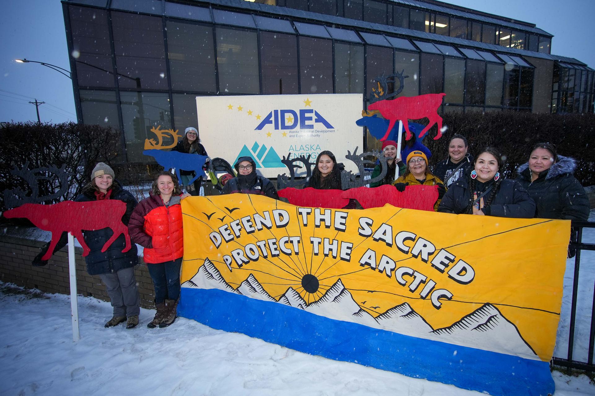 Ten people smile and look at the camera as light snow falls before sunrise. They are holing a very large sign that reads: DEFEND THE SACRED PROTECT THE ARCTIC. Behind the text is bright yellow, and a mountain landscape with blue water at the bottom. Some participants are also holding red and blue large cut outs of caribou. 