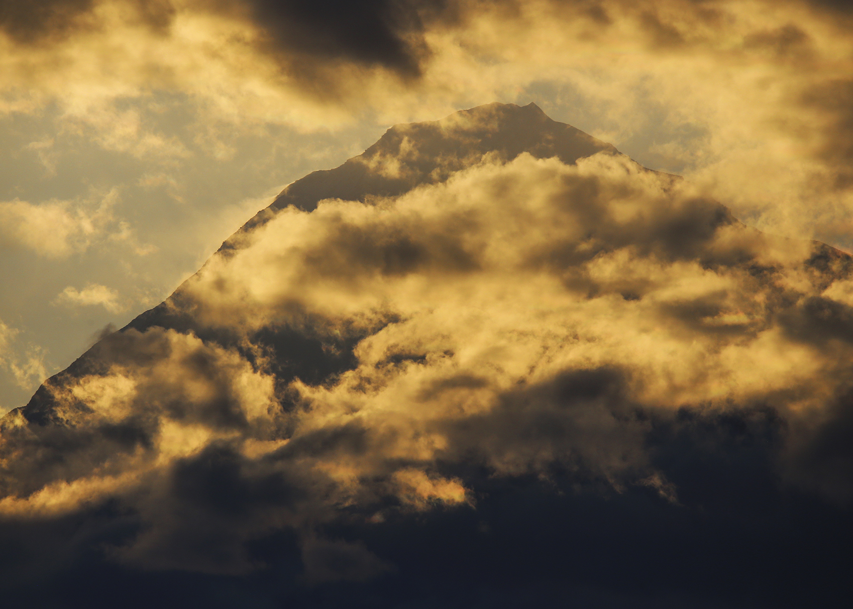 Yellow and dark grey clouds nearly cover Denali. The peak of the snowy mountain is barely visible in a gap in the clouds and the mountain looks shadowed. 