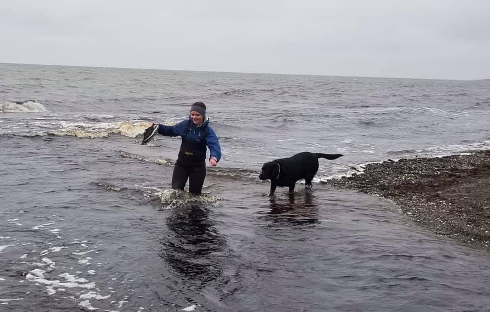 A woman holds her sneakers high above a body of water that she begins crossing. Next to her is a large black dog and the water is above the woman's knees. The sky is grey and behind her is the Kotzebue Sound, a large open body of water.