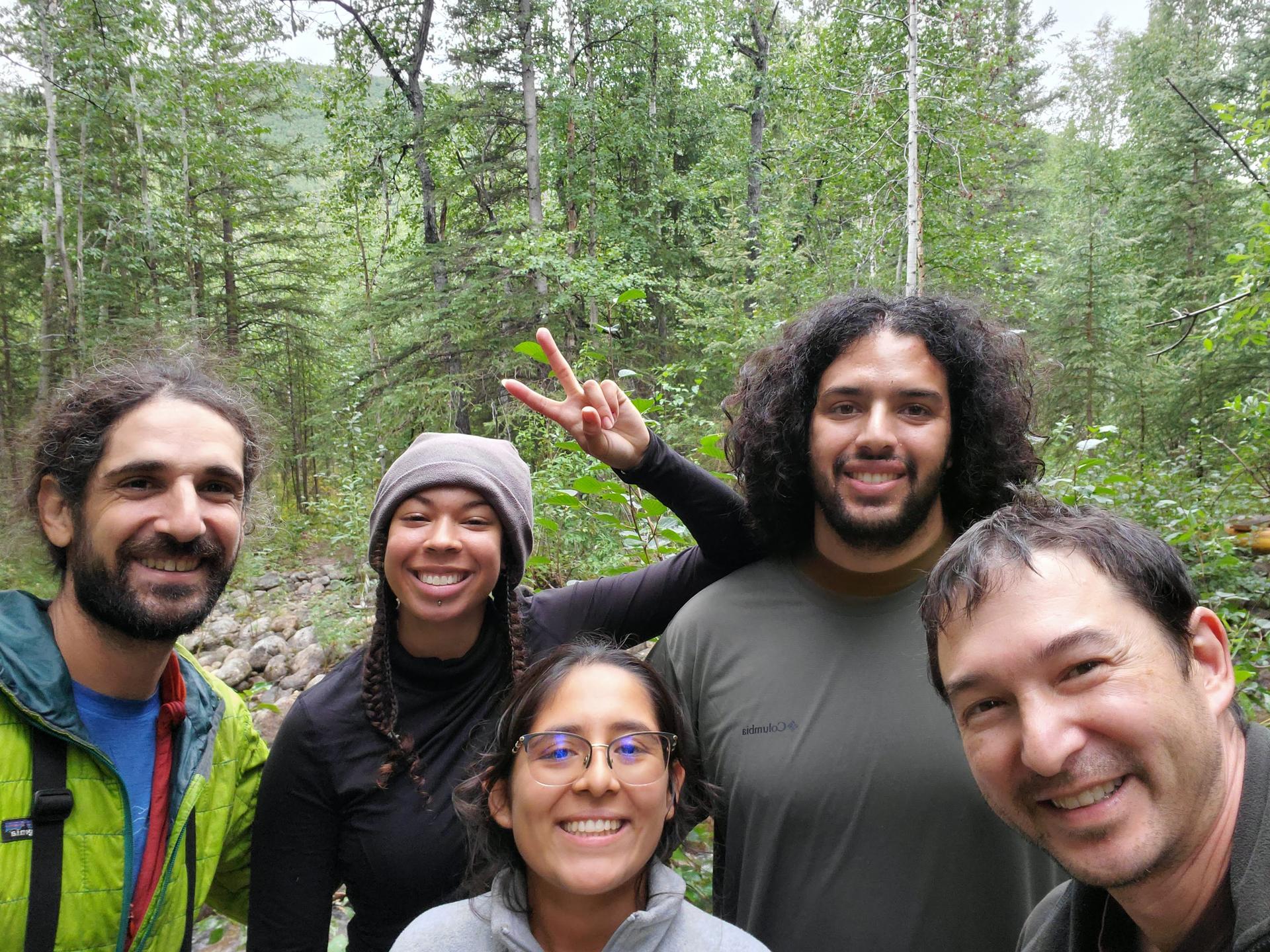 Against a green forest background, five people look at the camera and smile.