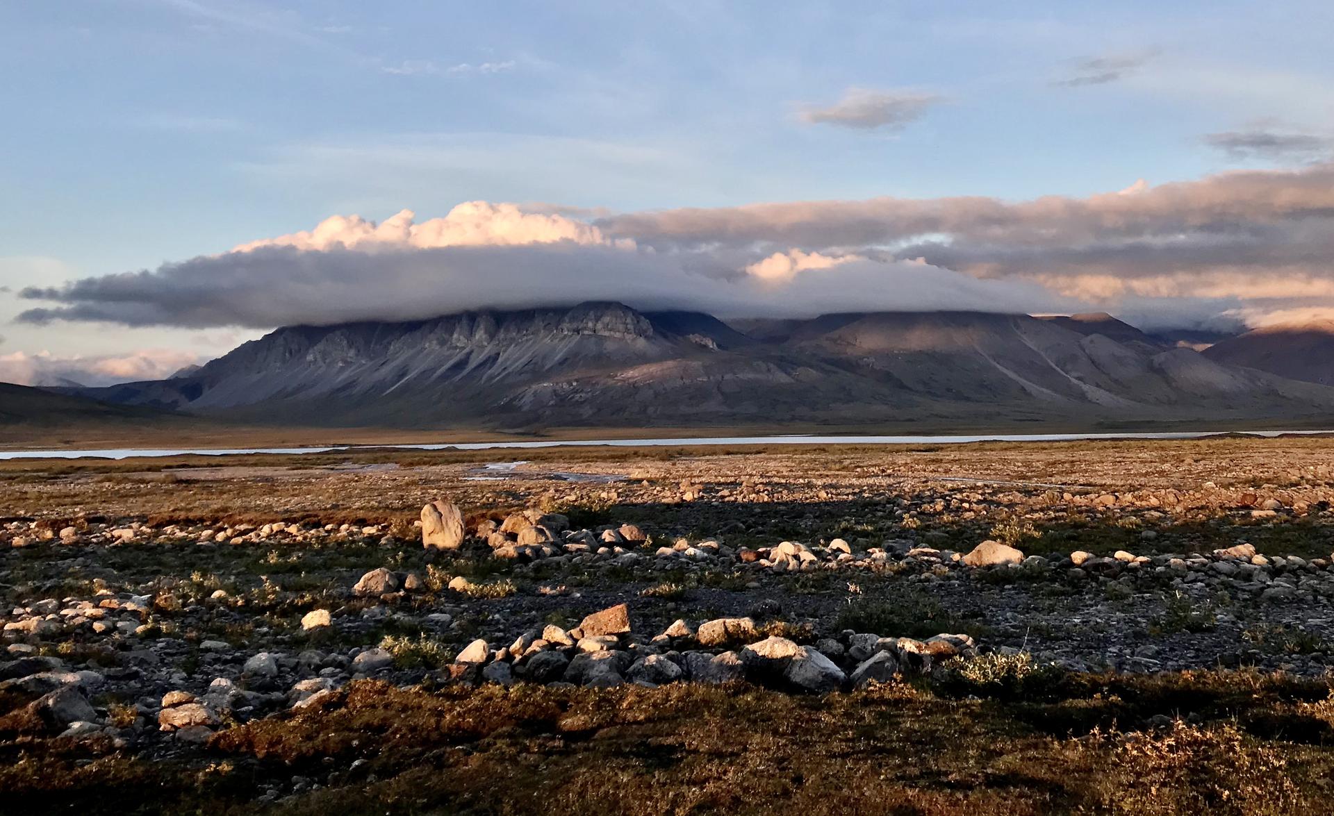 A landscape scene with warm hues on the rocky ground. In the distance, a thick layer of orange, grey, and purple clouds cover the rocky mountain peaks. Above is a soft blue sky.