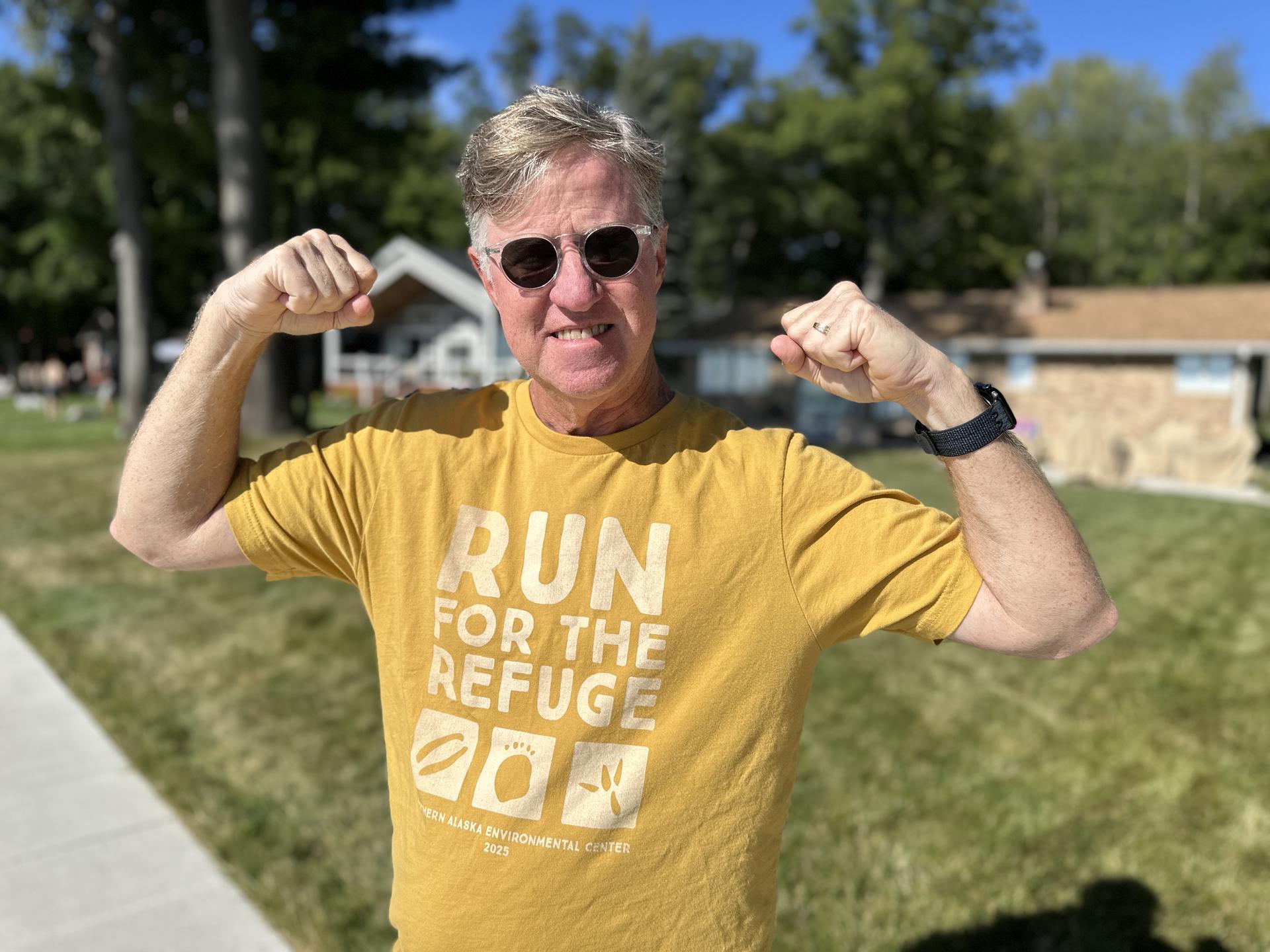 A man flexes his biceps and smiles at the camera while wearing a Run for the Refuge race t-shirt.