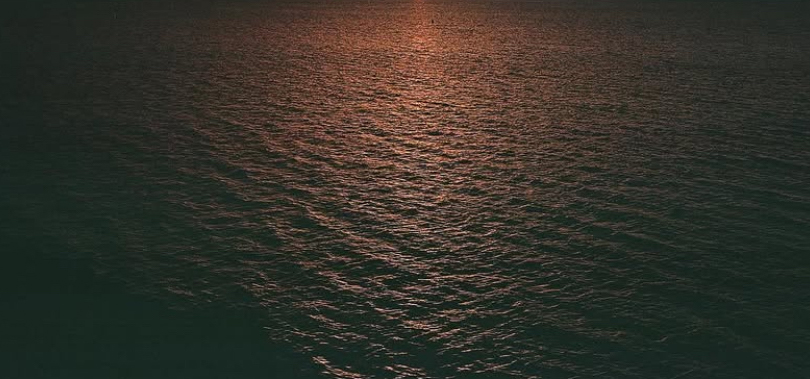 The setting sun casts an orange and pink hue on dark ocean water that encompasses the entire frame.