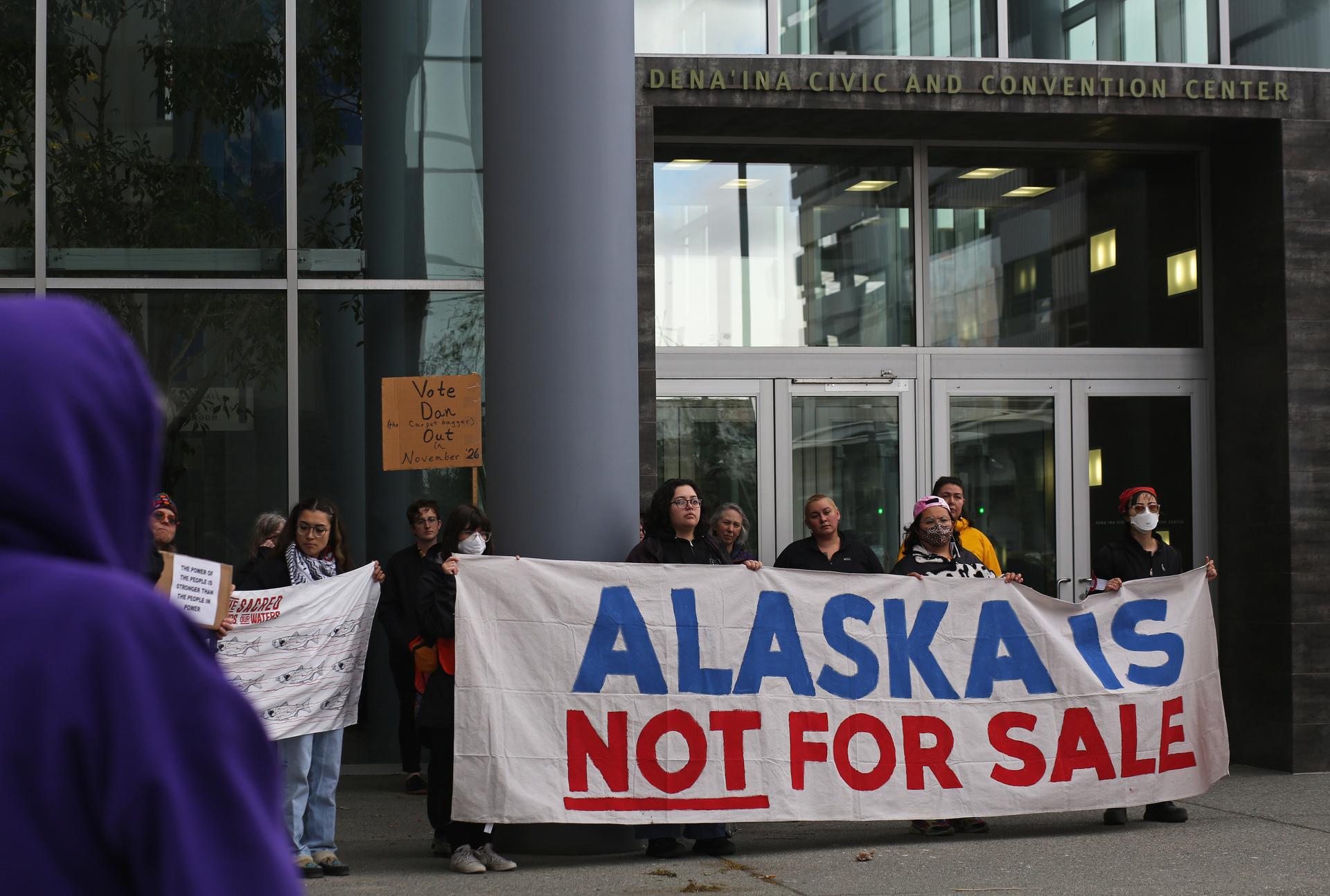 People gather outside a building in downtown Anchorage and protest a conference held by the governor of Alaska. Five people are predominately featured in this image and are holding a large cloth sign that says, “ALASKA IS NOT FOR SALE”