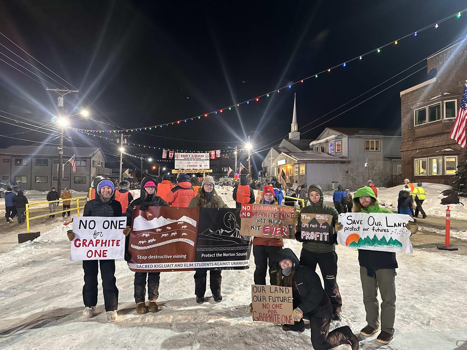 A group of seven people, layered in cold weather clothing hold signs demonstrating against mining proposals on a snow-covered street. Behind them, people linger around a banner marking the finish line of the Iditarod Trail. It's dark outside and street lights are shining into the camera.