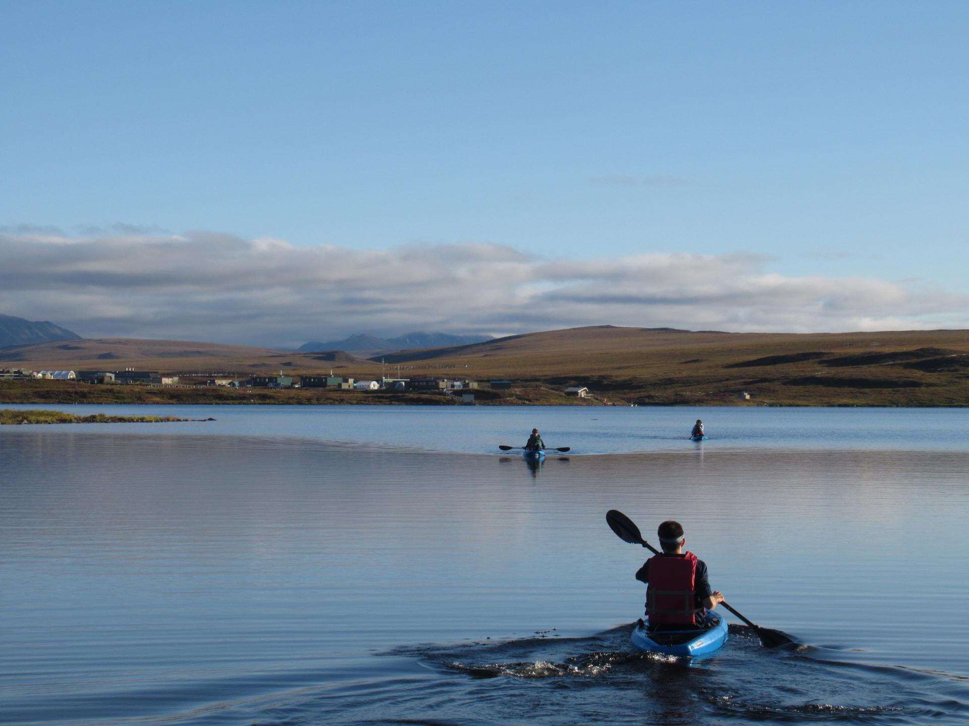 Three people paddle on a body of calm water. On the horizon is a hill covered in brown tundra.