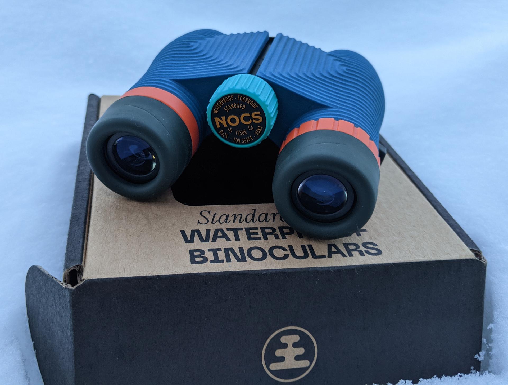 A colorful pair of binoculars (in navy blue, teal, and orange) rest on the NOCS-branded cardboard box in the snow.