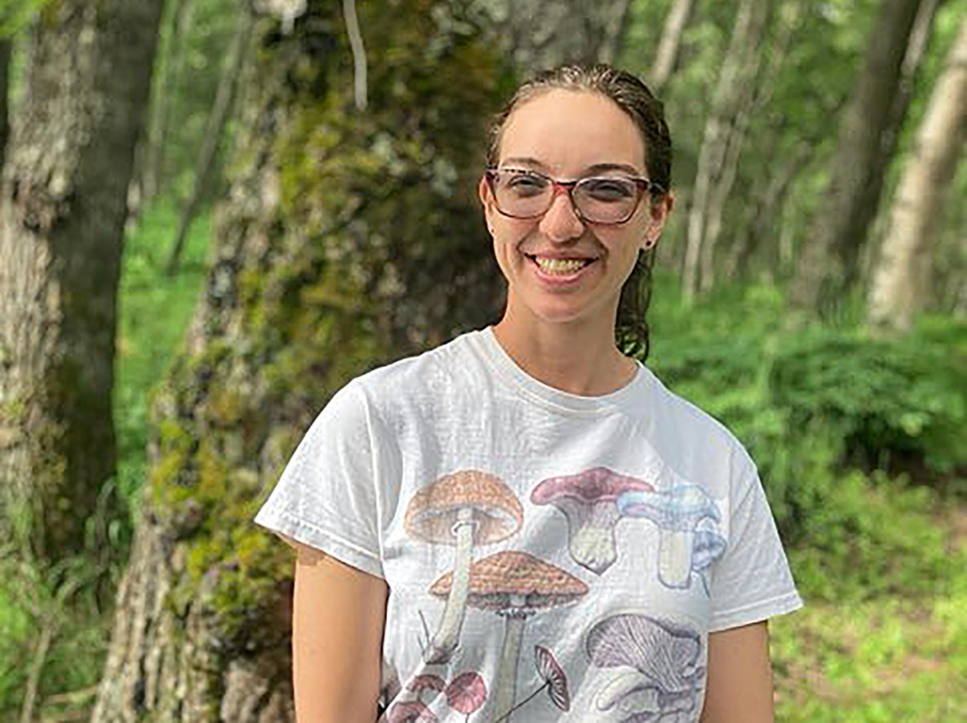 Christin Swearingen smiles at the camera as she stands in front of a large tree and is surrounded by a green wooded area. She is wearing a white t-shirt with mushrooms on it.