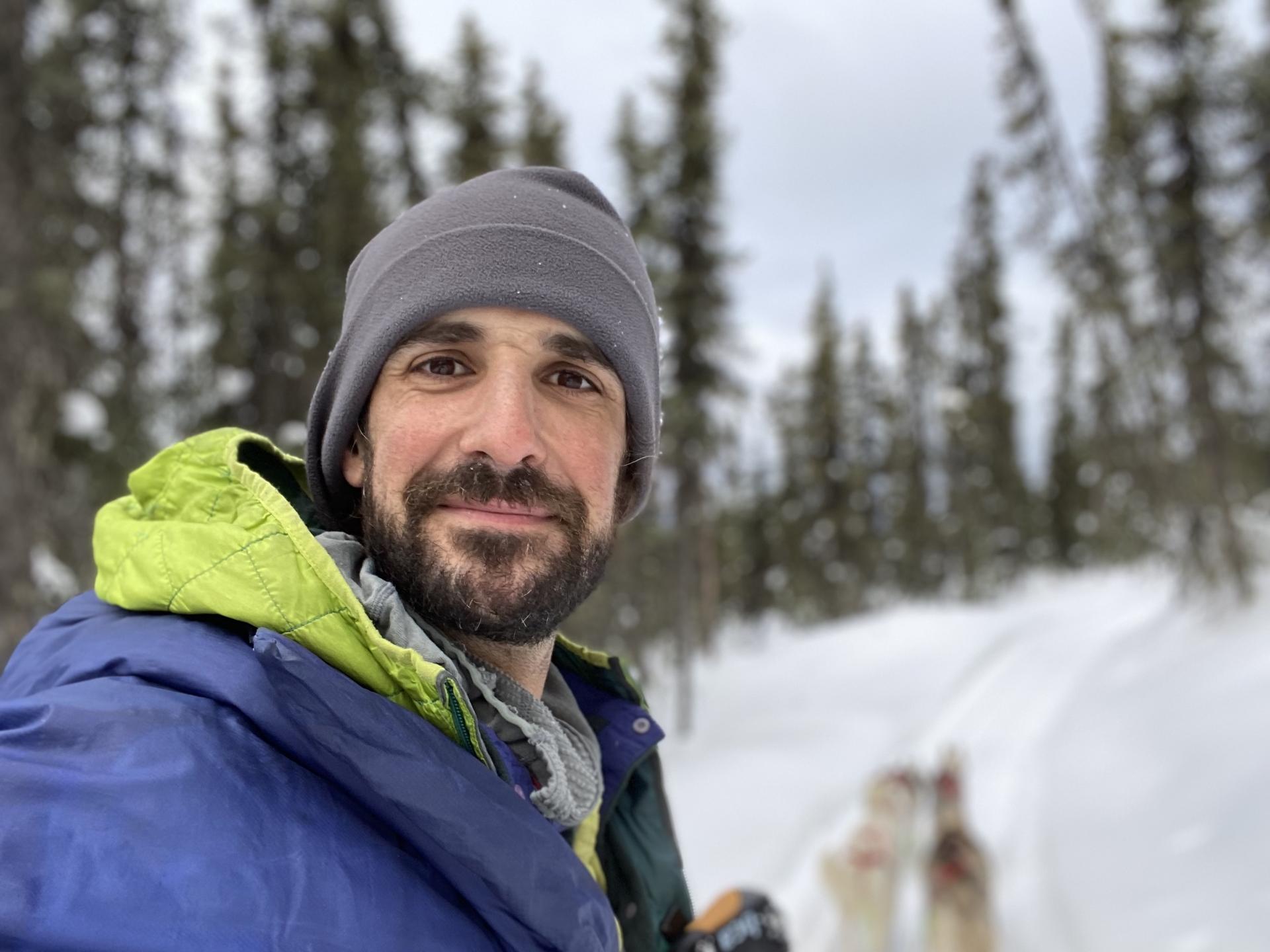 Ryan Arash Marsh looks to a camera he is holding and softly smiles. Behind him, out of focus, is a small dog team on a snowy trail surrounded by skinny spruce trees.