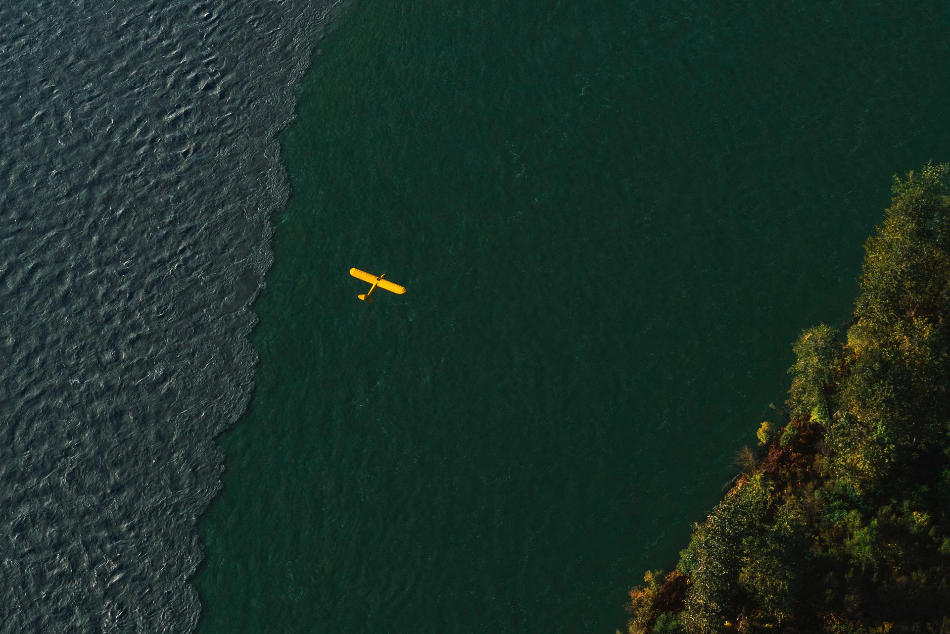 A bright yellow, small plane flies above the green/blue Susitna River. To the bottom right are green trees along the shoreline.