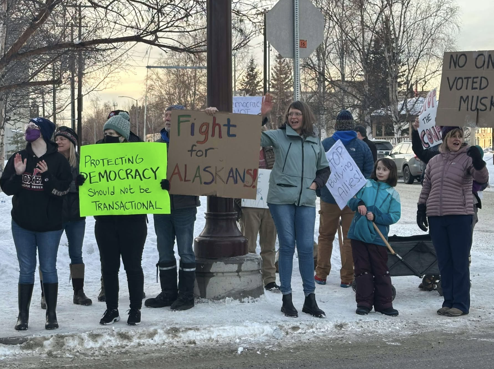 A crowd of people in winter gear stand on a snowy sidewalk by a road and protest the layoffs of federal employees. Signs read: ''Protecting democracy should not be transactional'', ''Fight for Alaskans'', and ''Stop bullying allies''.