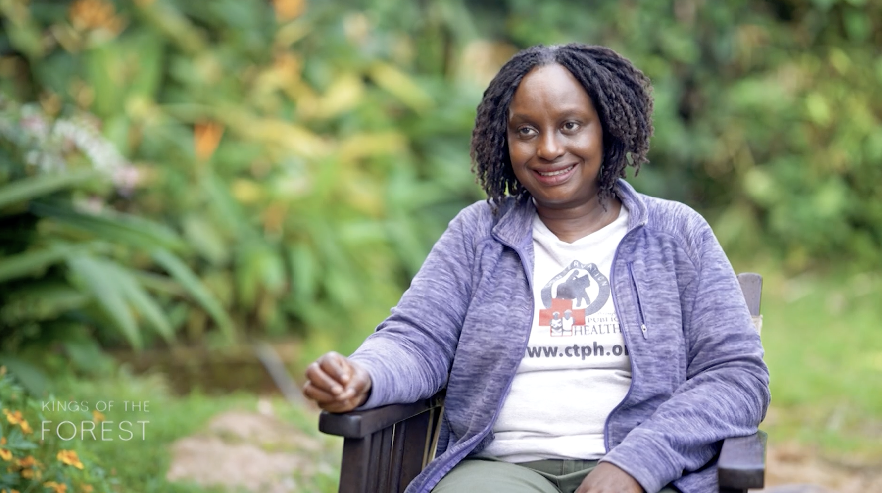 Dr. Gladys Kalema-Zikusoka, wearing a purple zip up sweater, Conservation Through Public Health shirt and sitting in an interview chair smiles at the interviewer in front of a bright green plant background.