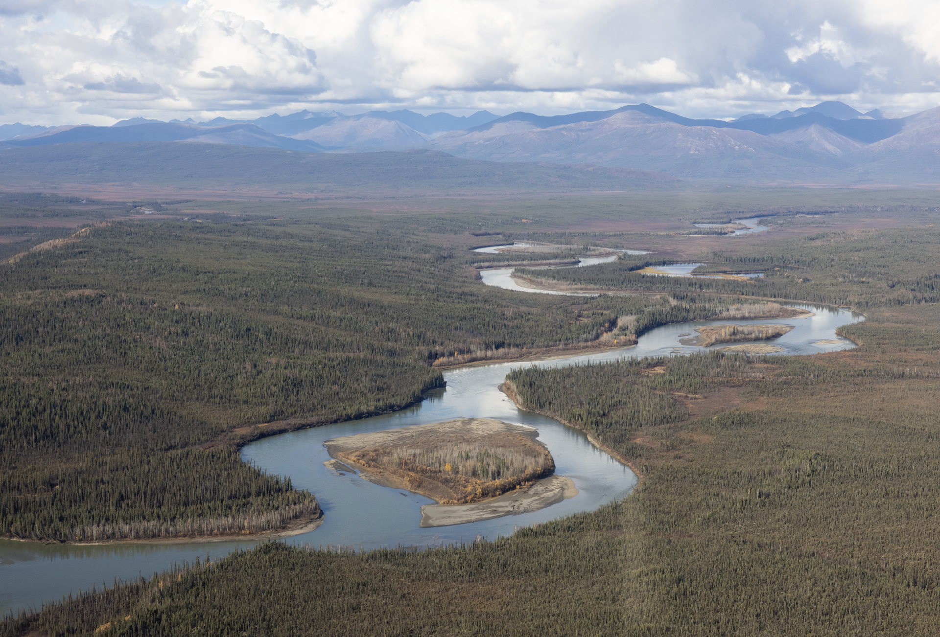 An aerial landscape image of a winding river, tundra, trees, with mountains on the horizon.