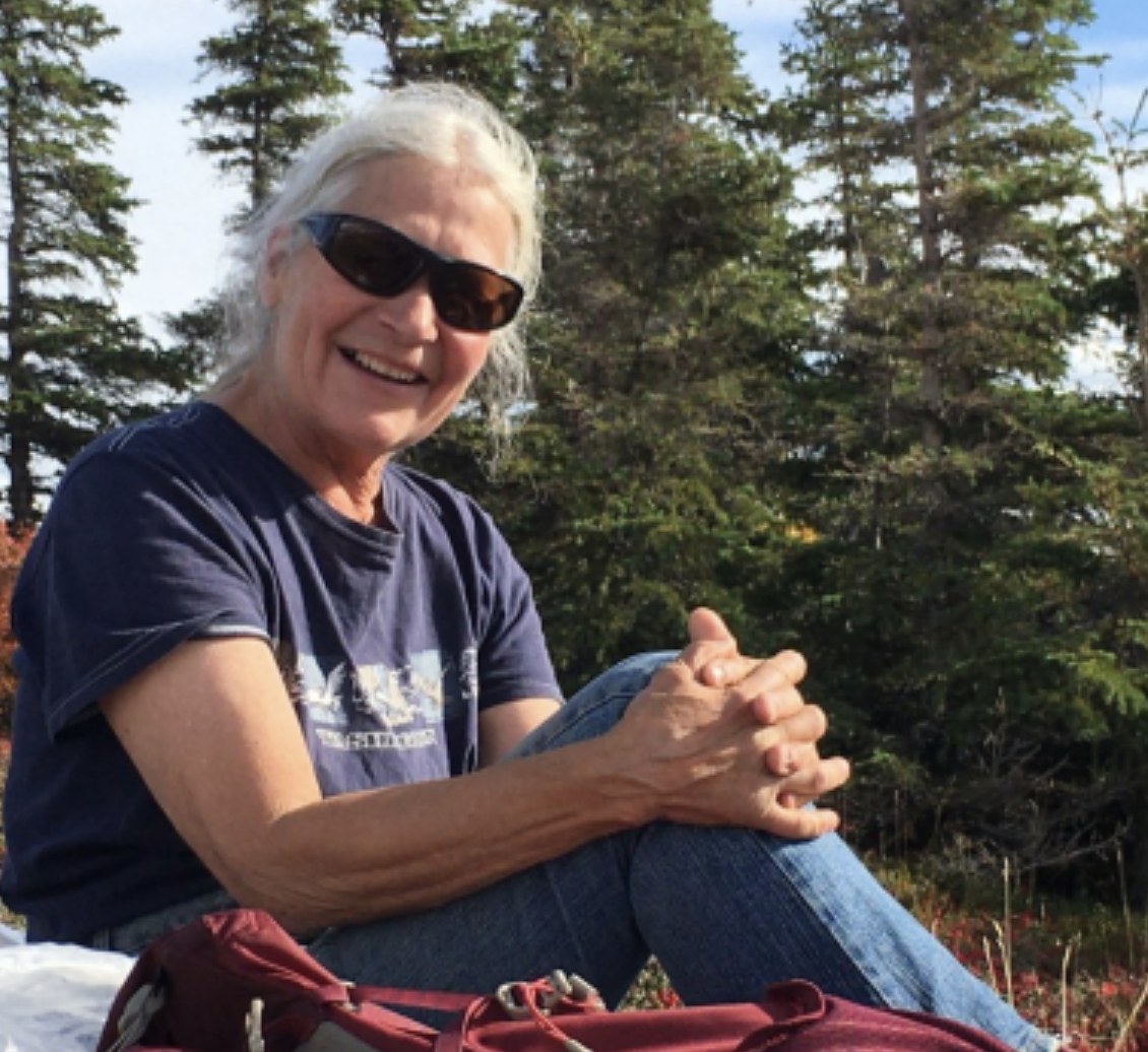 Karen Toland sits in the grass and interlaces her fingers together as she holds her knee. She's looking at the camera and smiling wearing sunglasses.