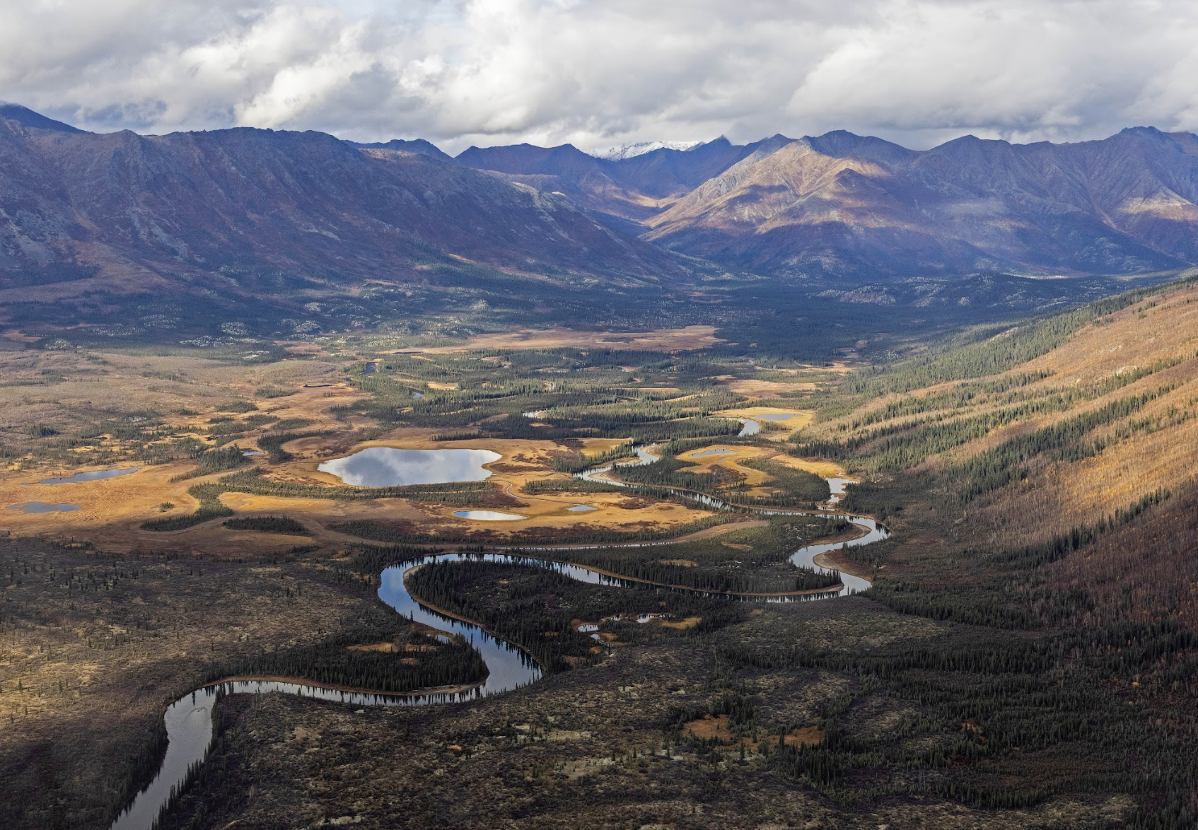 An aerial image showing a winding river through a mountainous valley surrounded by tundra and trees and ponds.