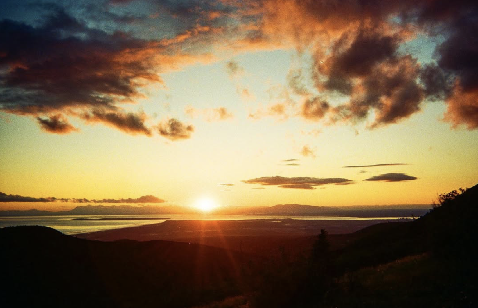 The sun sets and casts an orange and yellow hue to the sky. Clouds are highlighted peach and the foreground has hills in shadow.