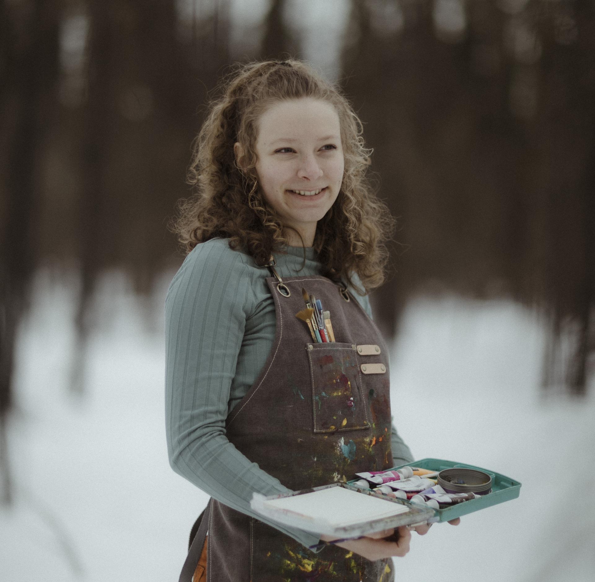 Tehya McLeod stands in a snowy forest with an artist's smock on that has colorful paint on it. She is holding a teal paint pallet and has paint brushes tucked into a pocket on the top of the brown smock. 