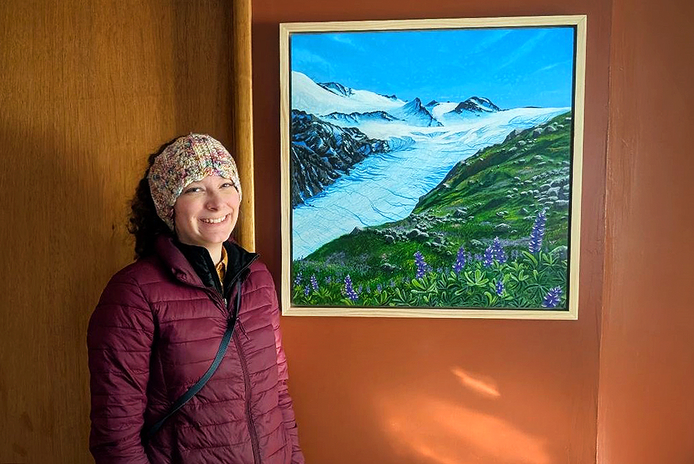 Tehya McLeod stands to the left of the frame, slightly in front of a large painting she created that shows a large glacier and mountainous icefield alongside bright green hills and purple lupine.