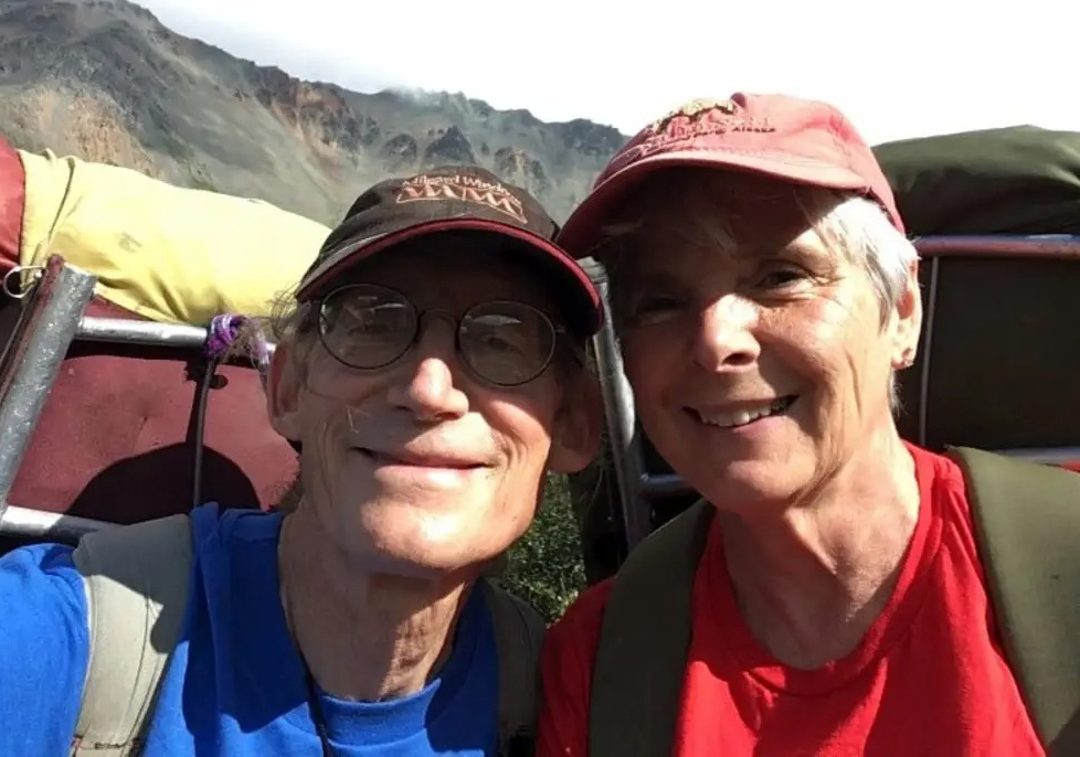 Wearing large framed backpacks, Stan (at left) and Barb (standing closely next to him) look to the camera that Stan is holding and smile. They are both wearing ball caps and there's a rocky mountainside behind them.
