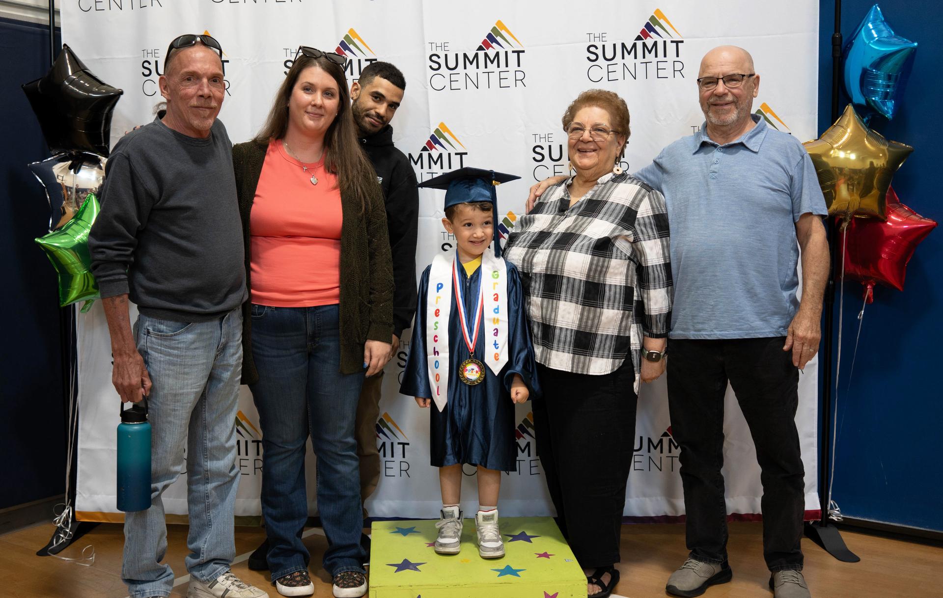 Family members pose with a young boy in preschool graduate robes in front of a white background with The Summit Center logo on it.
