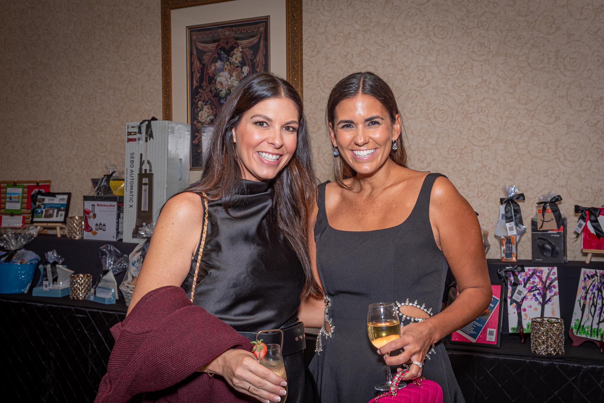Two women in elegant black dresses smile in front of a table of silent auction items.