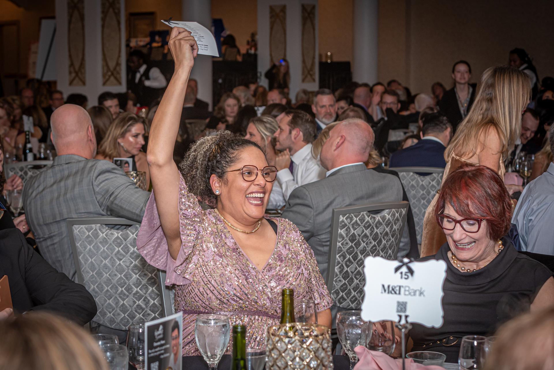 An elegantly dressed woman raises her hand to bid on an item at a gala function.