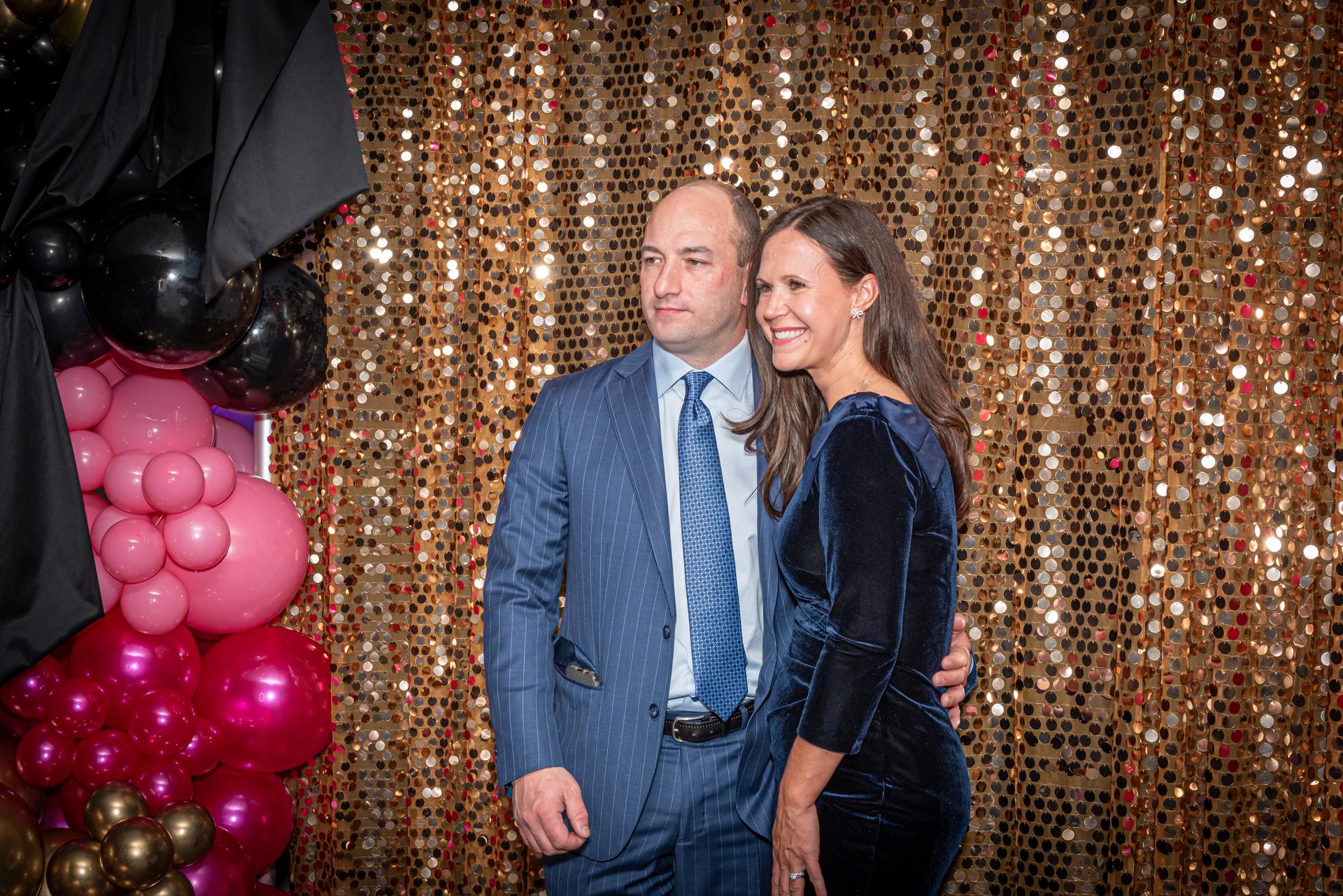 A couple stands in front of a gold shimmering backdrop.