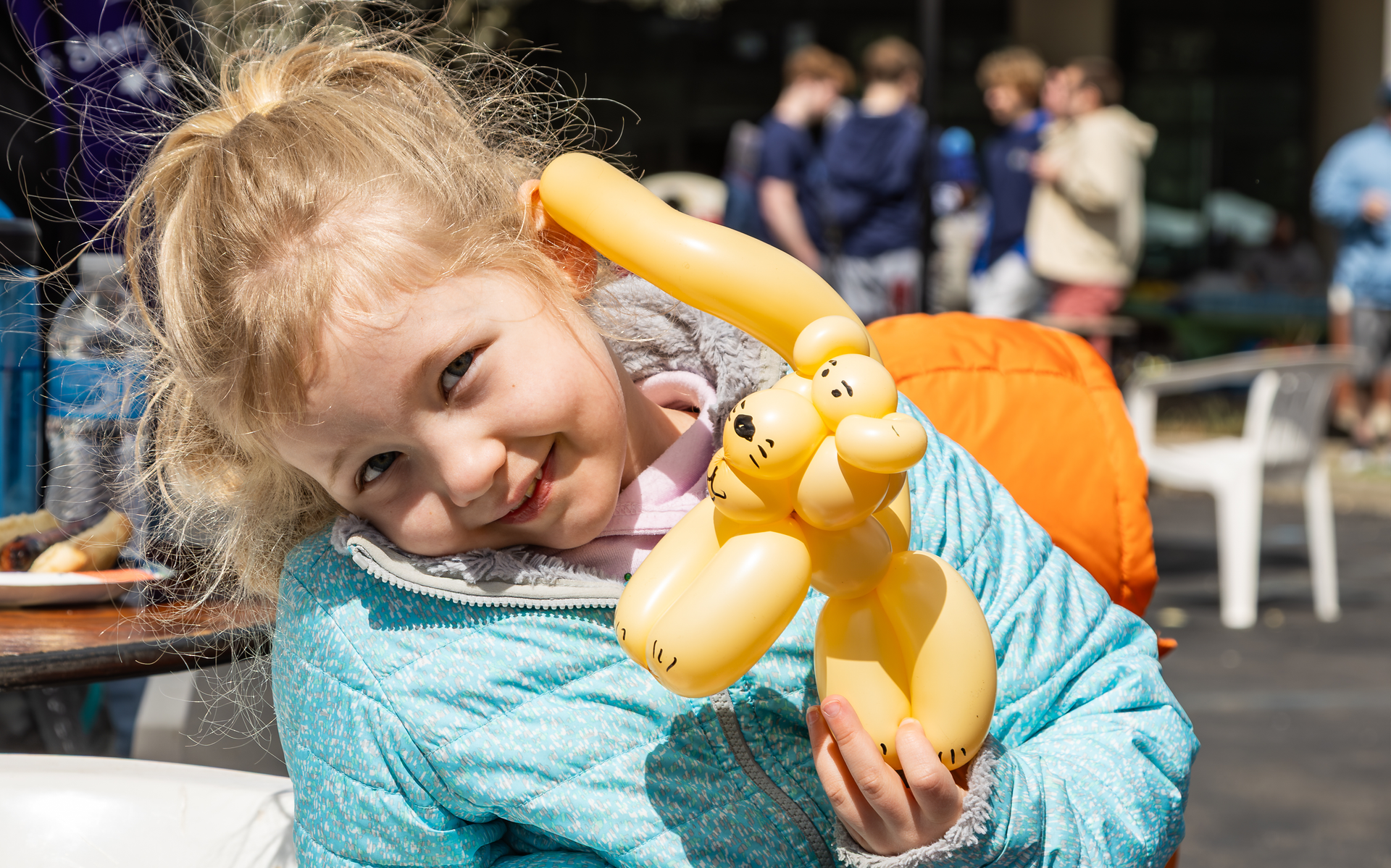 A young girl in a blue jacket smiles as she holds a yellow balloon animal.