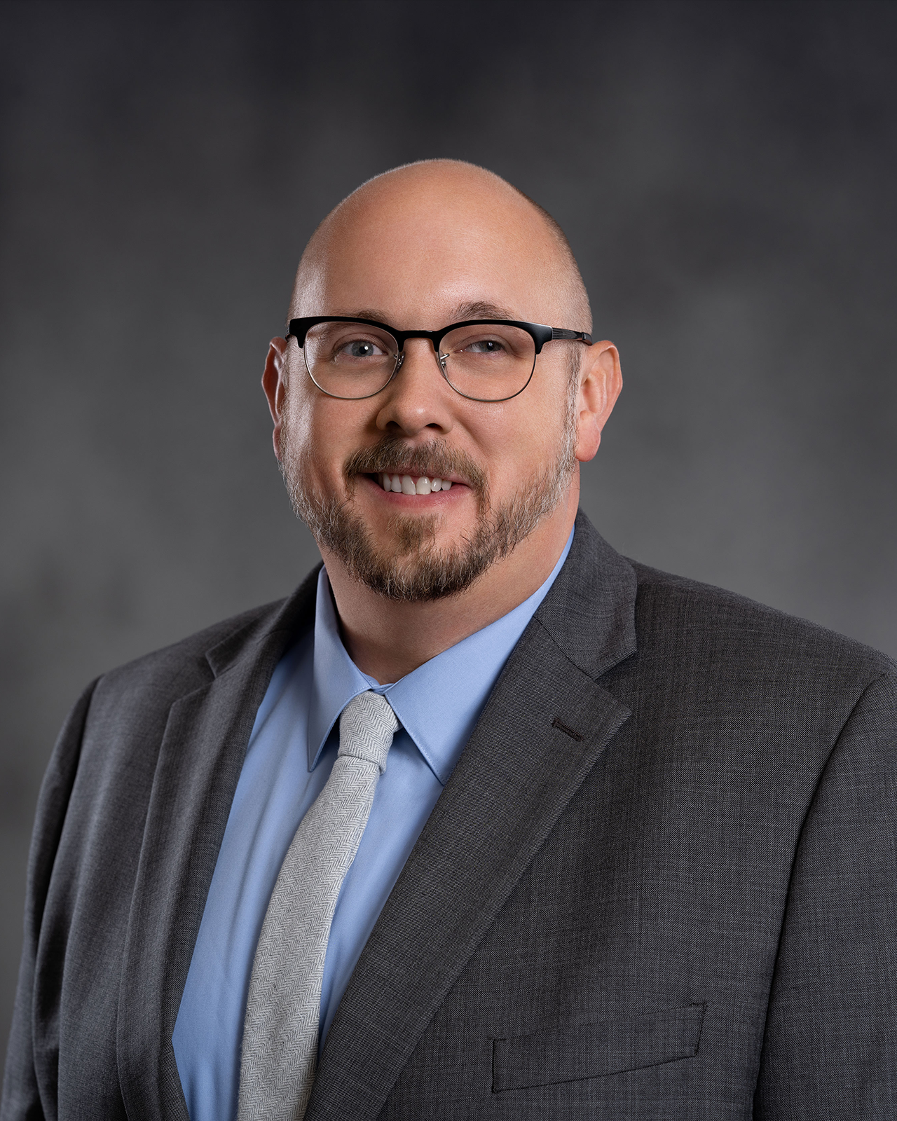 A man wearing glasses wearing a gray suit with a light blue shirt and light gray tie in front of a gray background