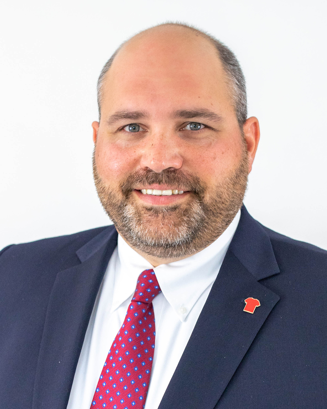 A man with a navy blue suit, white shirt, and red tie in front of a white background