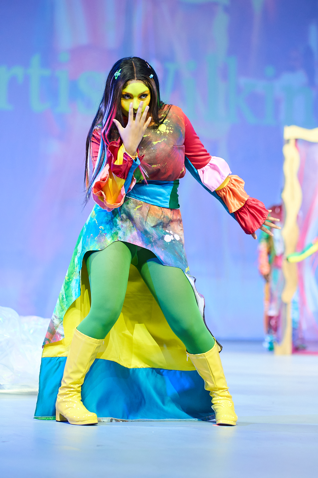 A woman with a yellow painted face and wearing rainbow colored clothing holds her hand up to her mouth on a runway.