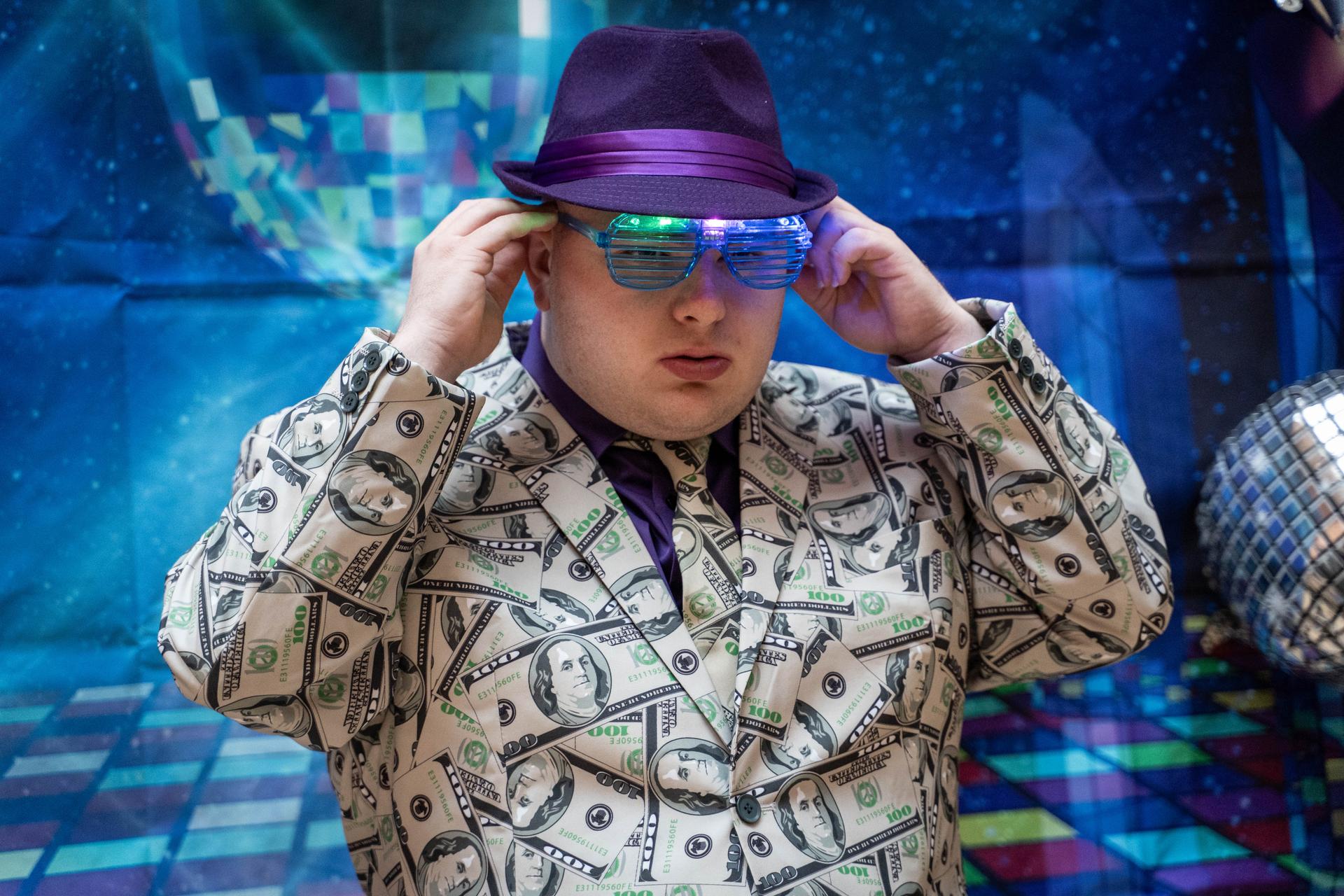 A young man wearing a dollar bill suit jacket and purple hat puts on blue stunner shades.