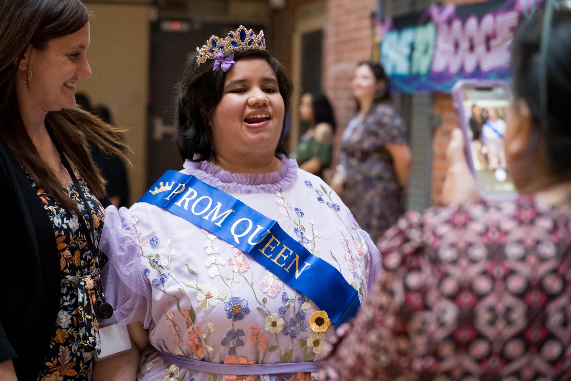 A young woman wearing a floral dress, crown, and prom queen sash smiles.