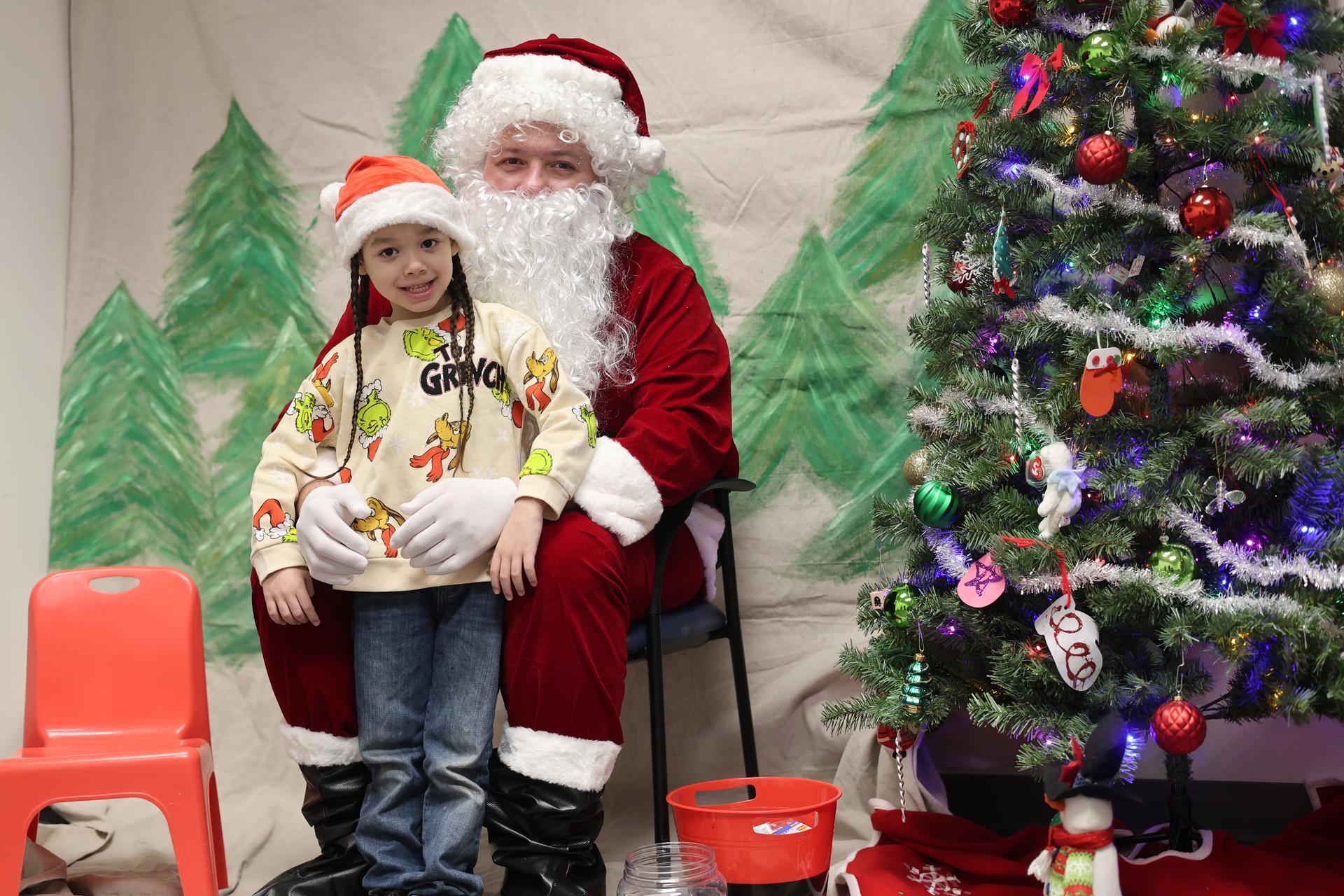 A young child in a Grinch sweater poses with Santa next to a decorated tree.