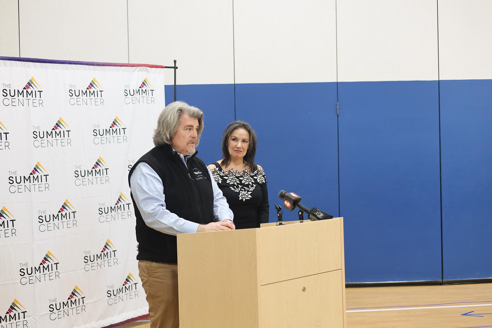 A man and woman dressed professionally stand in front of a podium with a microphone in a gymnasium.