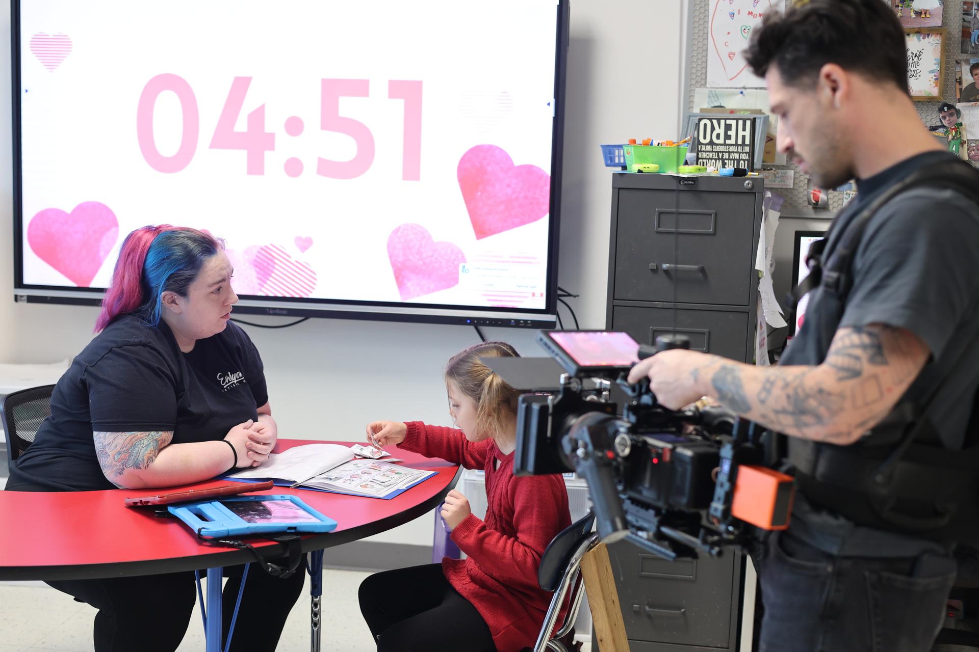A man in a gray shirt films a teacher and her student at a desk.