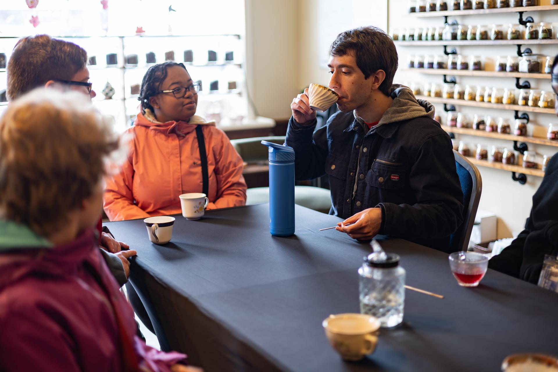 A young man takes a sip of tea while sitting at a table with friends.