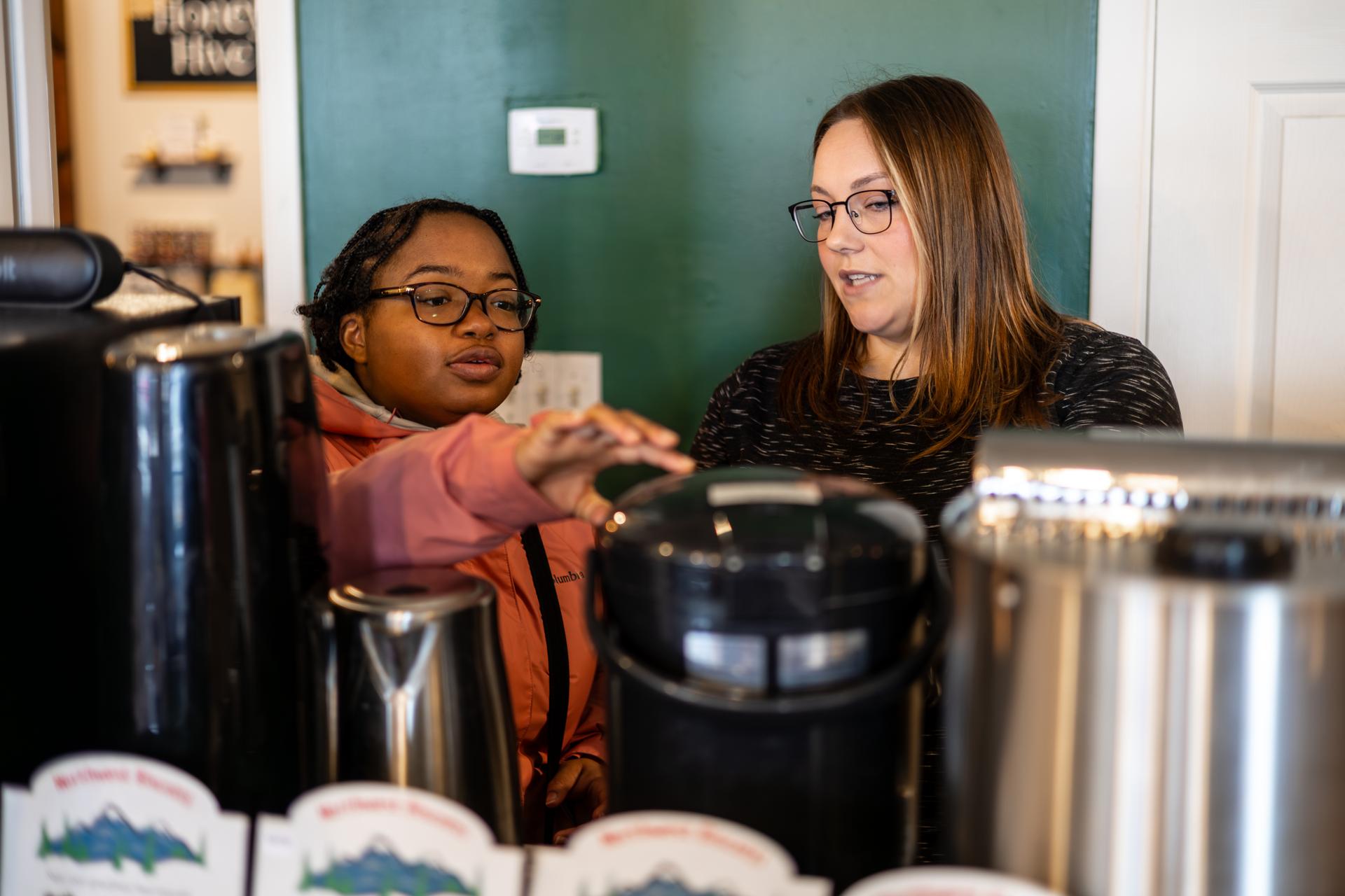 Two young women use tea brewing appliances.
