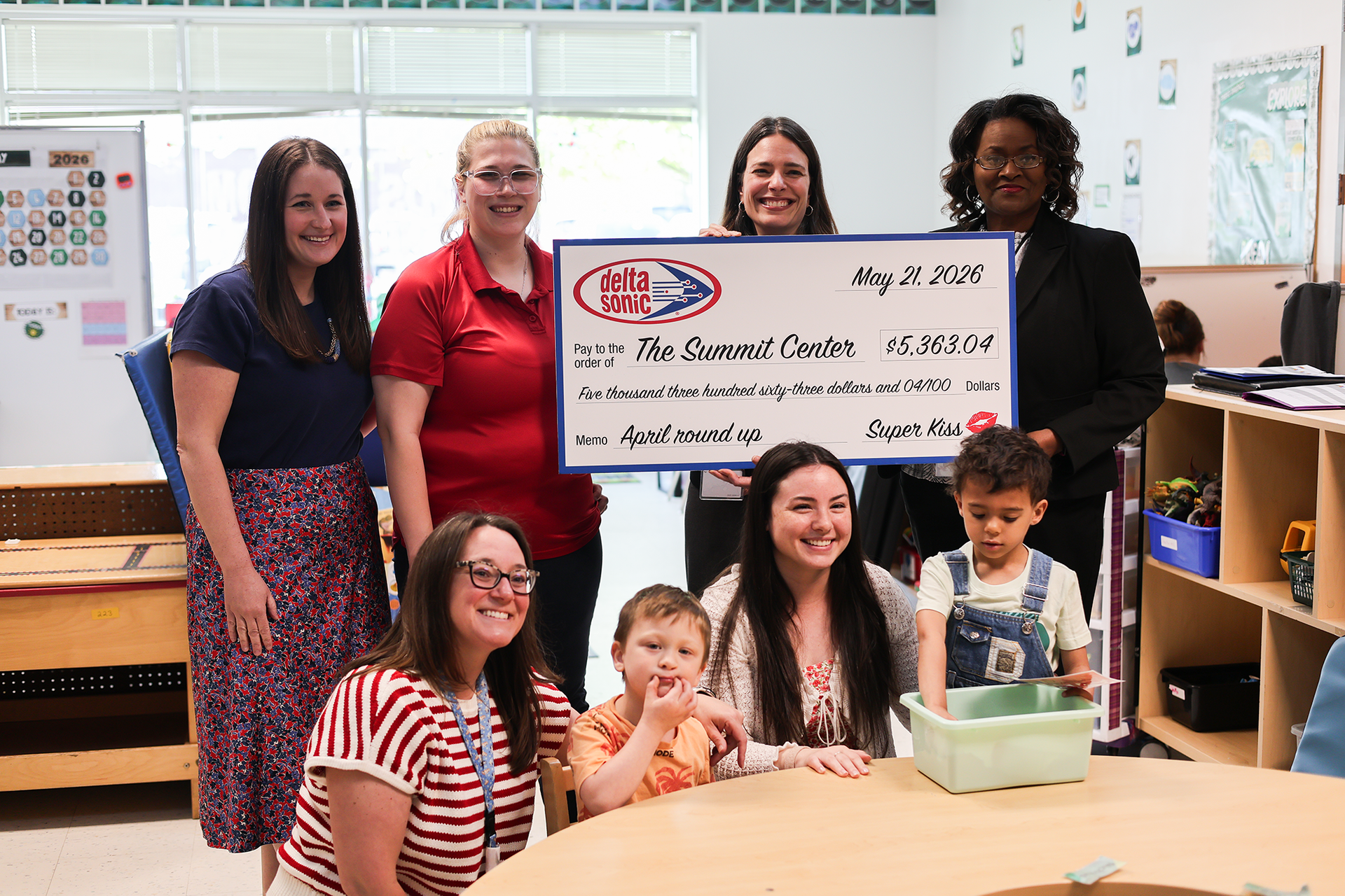 A group of people and two children stand around a table in a classroom holding a check from Delta Sonic.