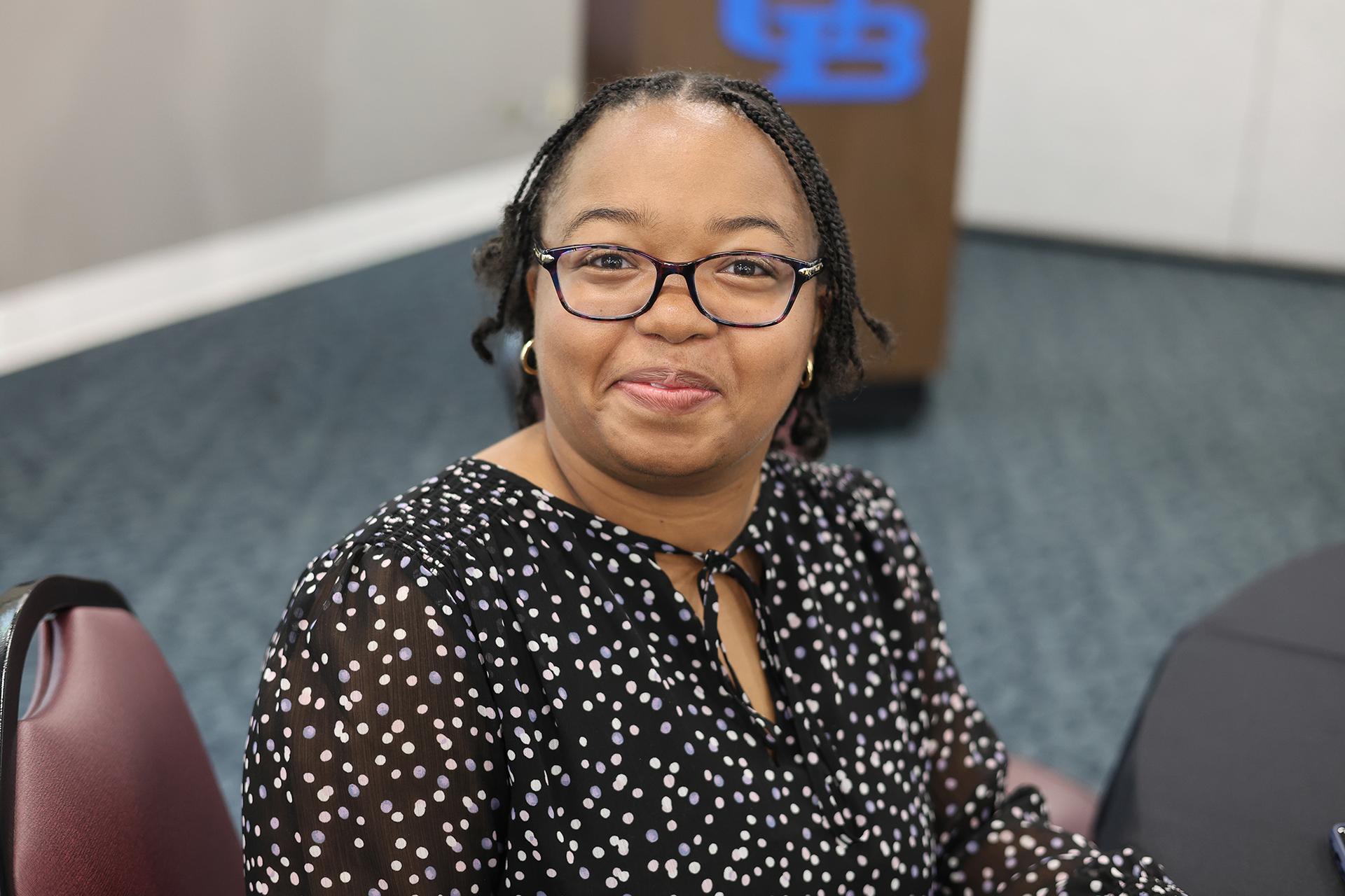 A young woman wearing a black dress and sitting in a conference room smiles.